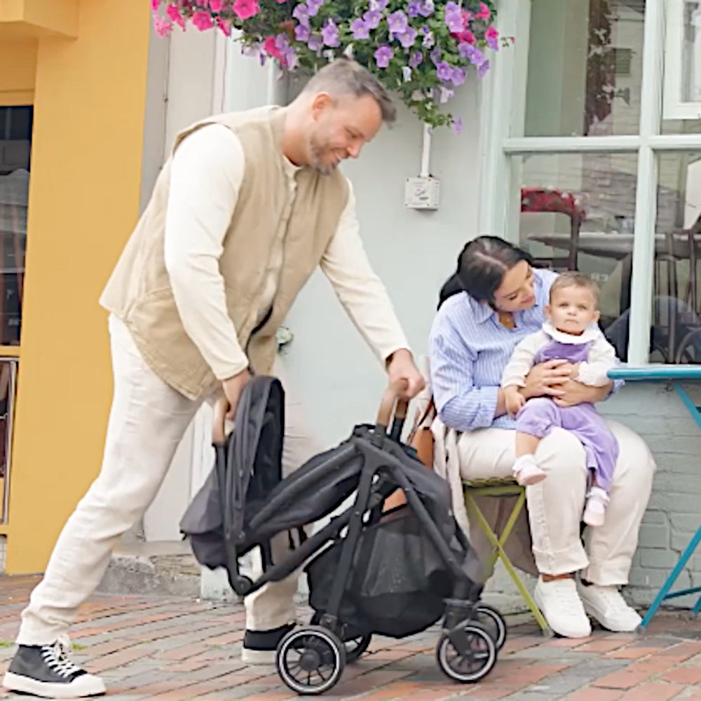 Dad unfolding Black Joie Pact Pro lightweight compact pushchair, mum holding baby
