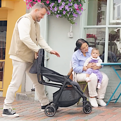 Dad unfolding Black Joie Pact Pro lightweight compact pushchair, mum holding baby